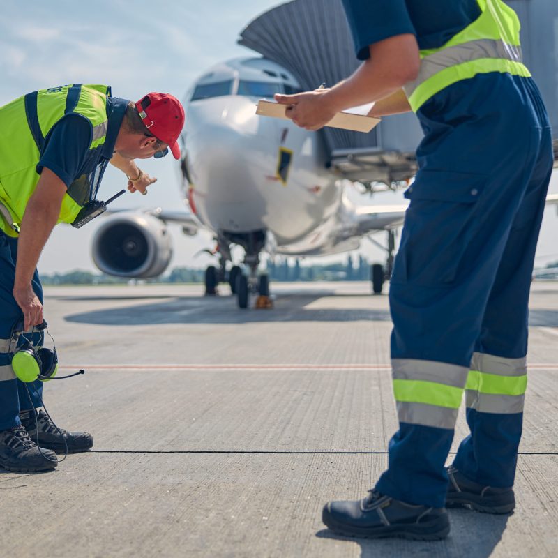 Airport worker dressed in uniform pointing at the parked plane to his Caucasian male colleague